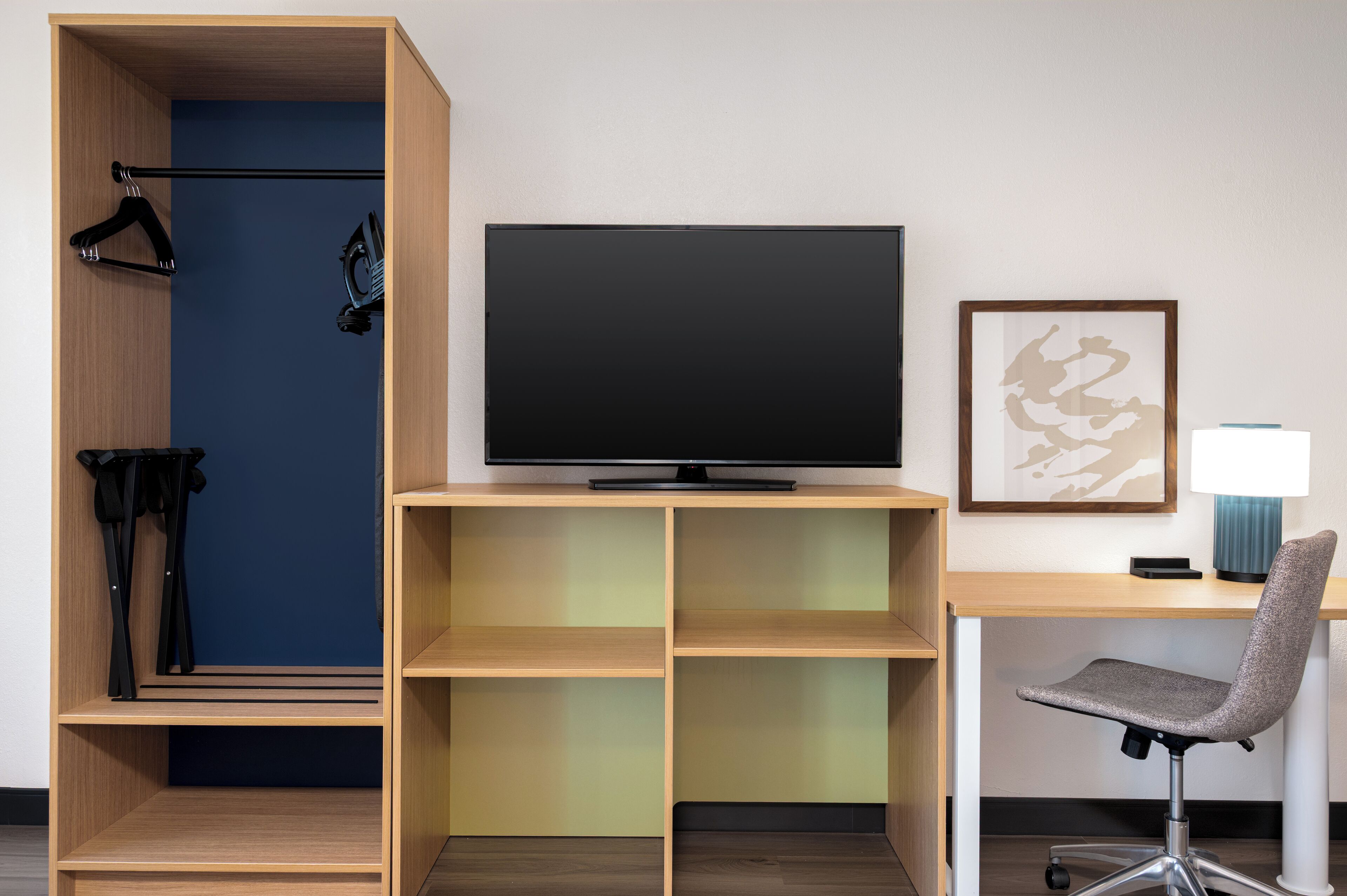 A hotel room with a TV, desk, chair, and a wardrobe with hanging clothes.