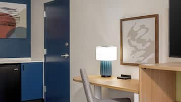 Habitación de hotel con escritorio, silla y lámpara. Se observa una puerta y un refrigerador pequeño.