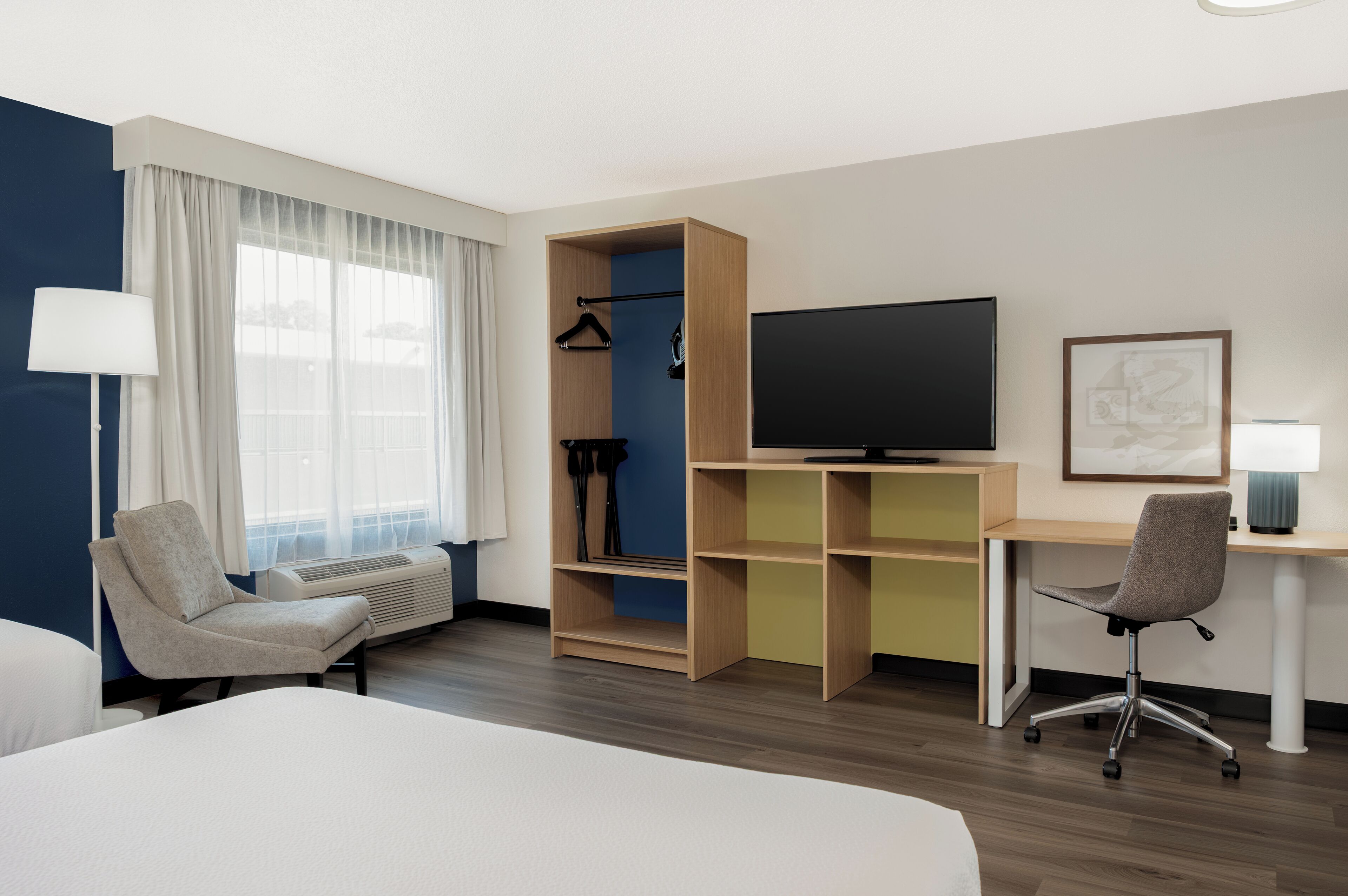 Une chambre d’hôtel comprenant un lit, un bureau, une chaise, une télévision, une armoire et une fenêtre avec des rideaux.