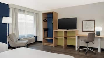 Une chambre d’hôtel comprenant un lit, un bureau, une chaise, une télévision, une armoire et une fenêtre avec des rideaux.