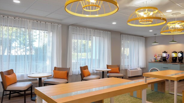 A modern hotel lobby with wooden tables and chairs, large windows with white curtains, and multiple pendant lights.