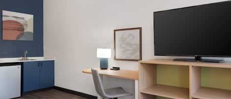 Una cucina moderna con lavastoviglie, mobili azzurri, una scrivania in legno, una sedia da ufficio grigia, una TV a schermo piatto e un quadro appeso al muro.