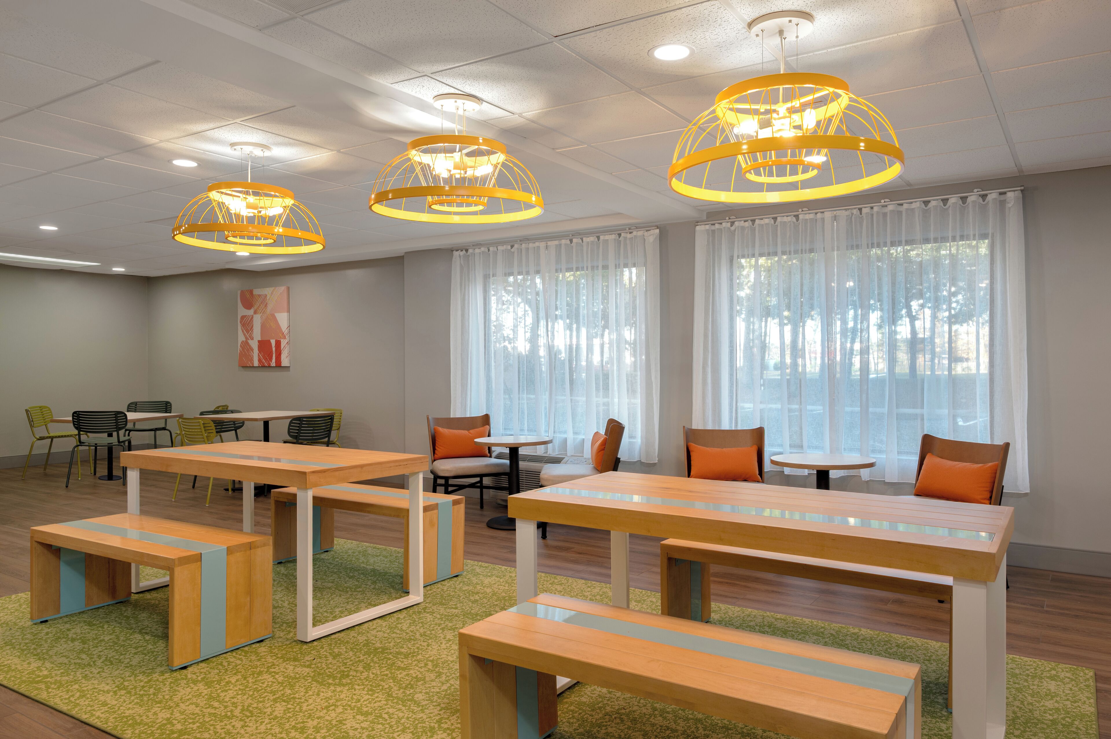 A modern meeting room with wooden tables and benches, large windows with sheer curtains, and a ceiling with recessed lighting and decorative yellow fixtures.