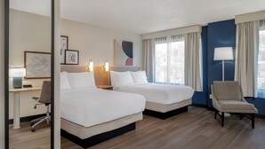 A hotel room with two beds, a desk with a chair, a lamp, and artwork on the wall.