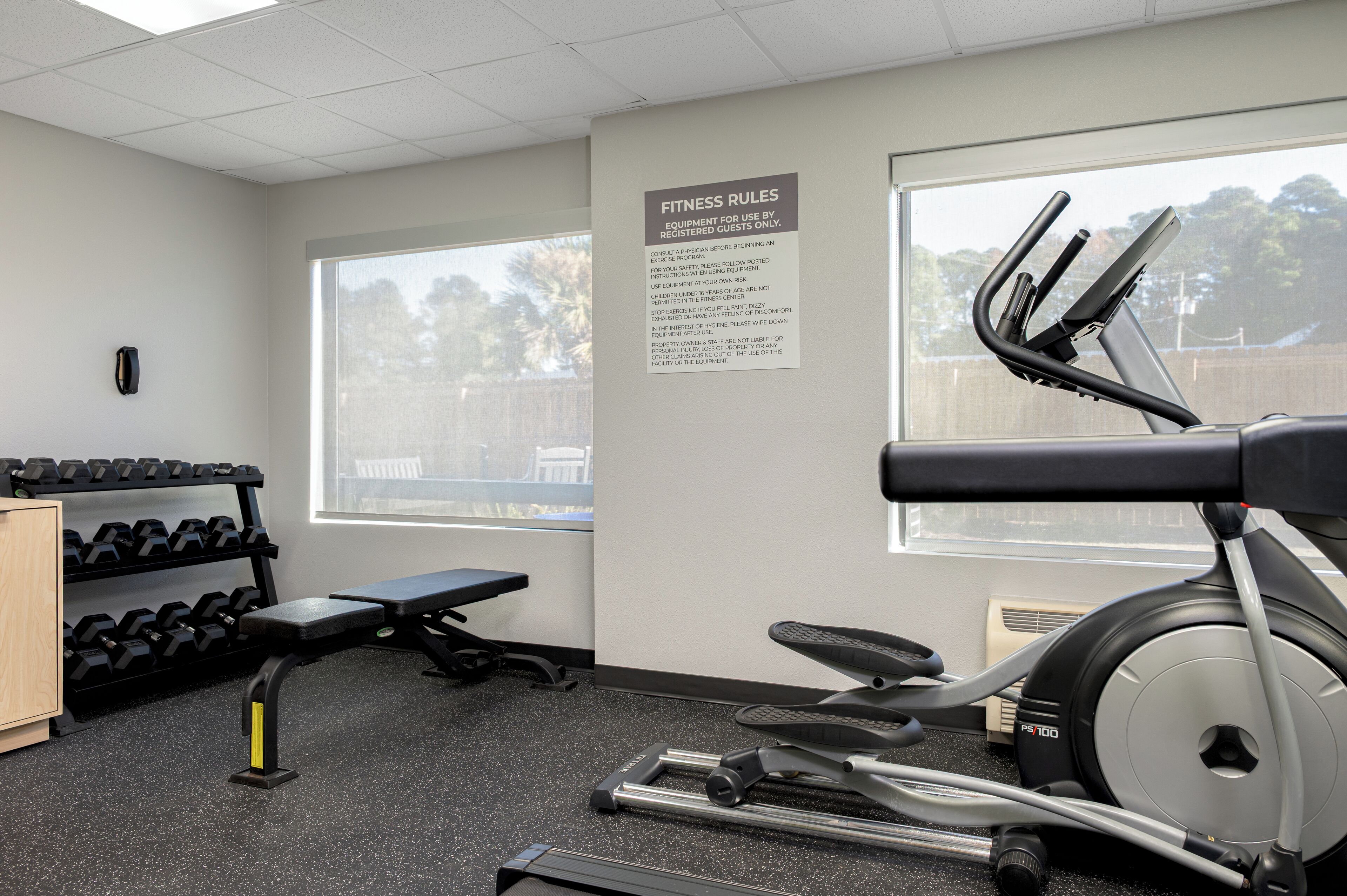 A gym with exercise equipment, a wall-mounted sign with fitness rules, and a window with a view of trees.