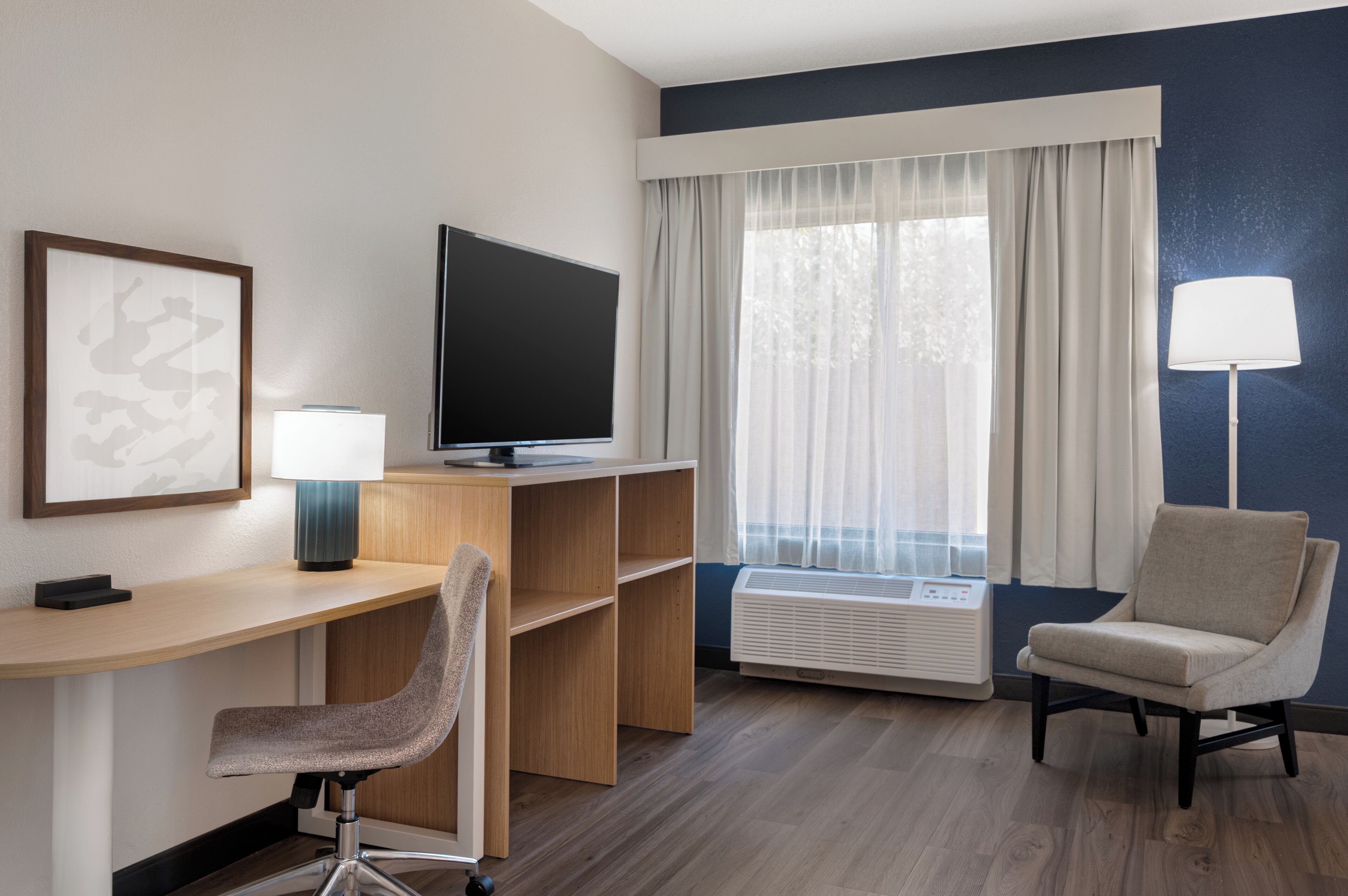 A hotel room with a desk, a chair, a television, a lamp, and a window with curtains.