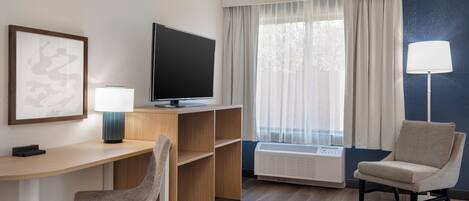 Habitación de hotel con escritorio, silla, televisión, lámpara y ventana con cortinas.