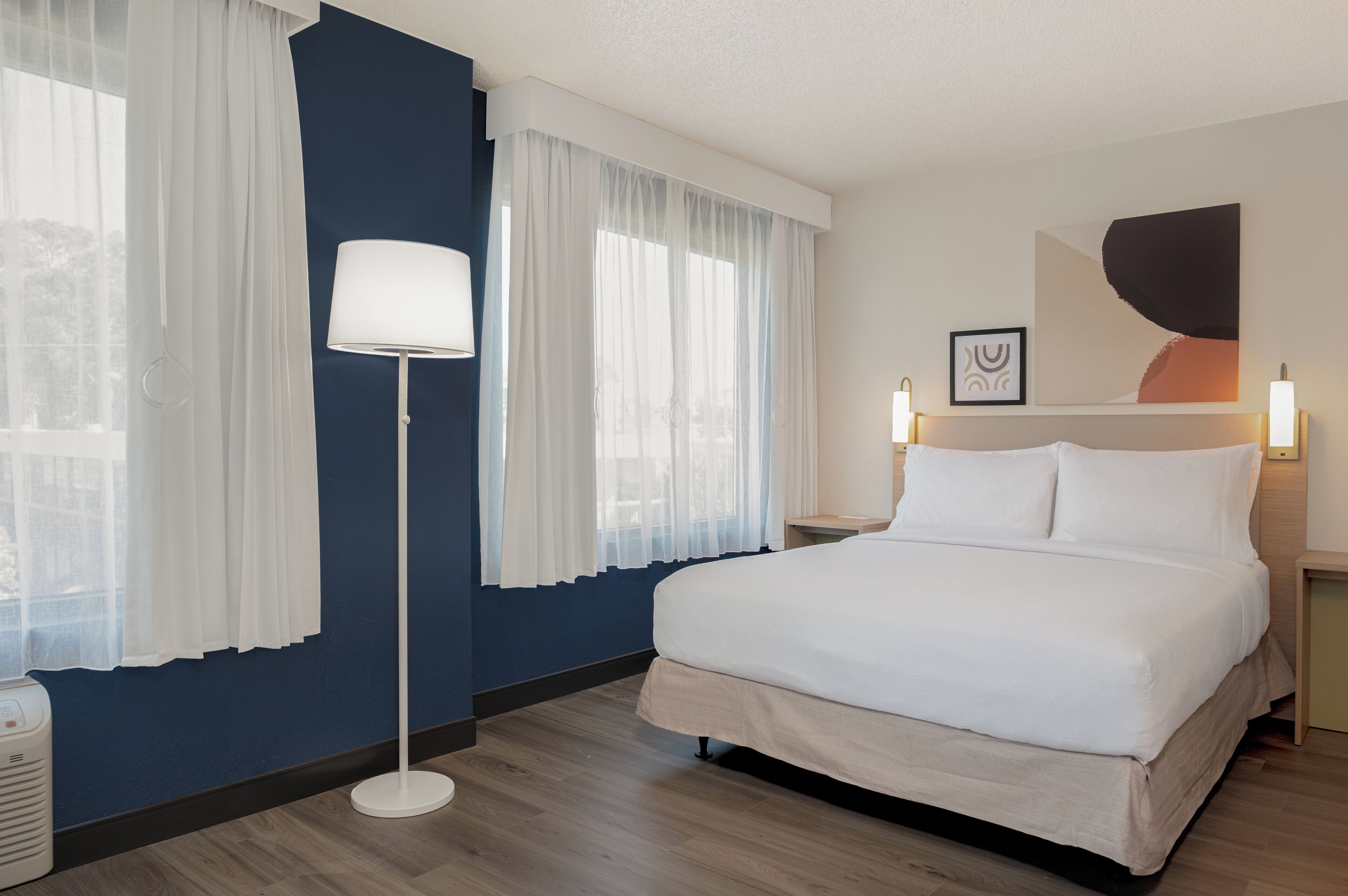 Ein Hotelzimmer mit Bett, Stehlampe, Nachttisch und einem großen Fenster mit durchsichtigen Vorhängen.