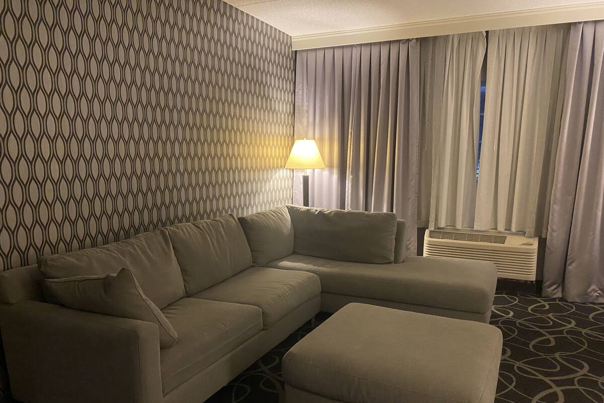 A modern hotel room with a sectional sofa, an ottoman, a patterned wall, a lamp, and a window with curtains.