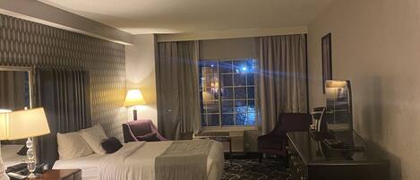 A hotel room with two beds, a desk, a chair, a lamp, a window with curtains, and a patterned carpet.