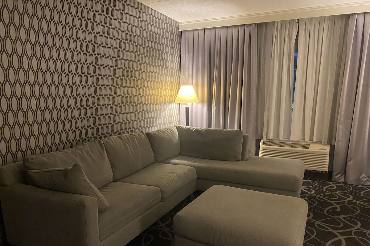 Une chambre d’hôtel avec un canapé d’angle, un pouf, un mur à motifs, une lampe, des rideaux et un appareil de climatisation.