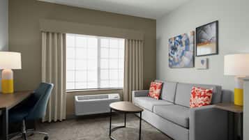 A hotel room with a grey sofa, a blue chair, a small round table, a standing heater, and a window with curtains.