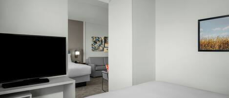 Habitación de hotel con un televisor de pantalla plana grande, una cama, un sofá gris y un cuadro en la pared.