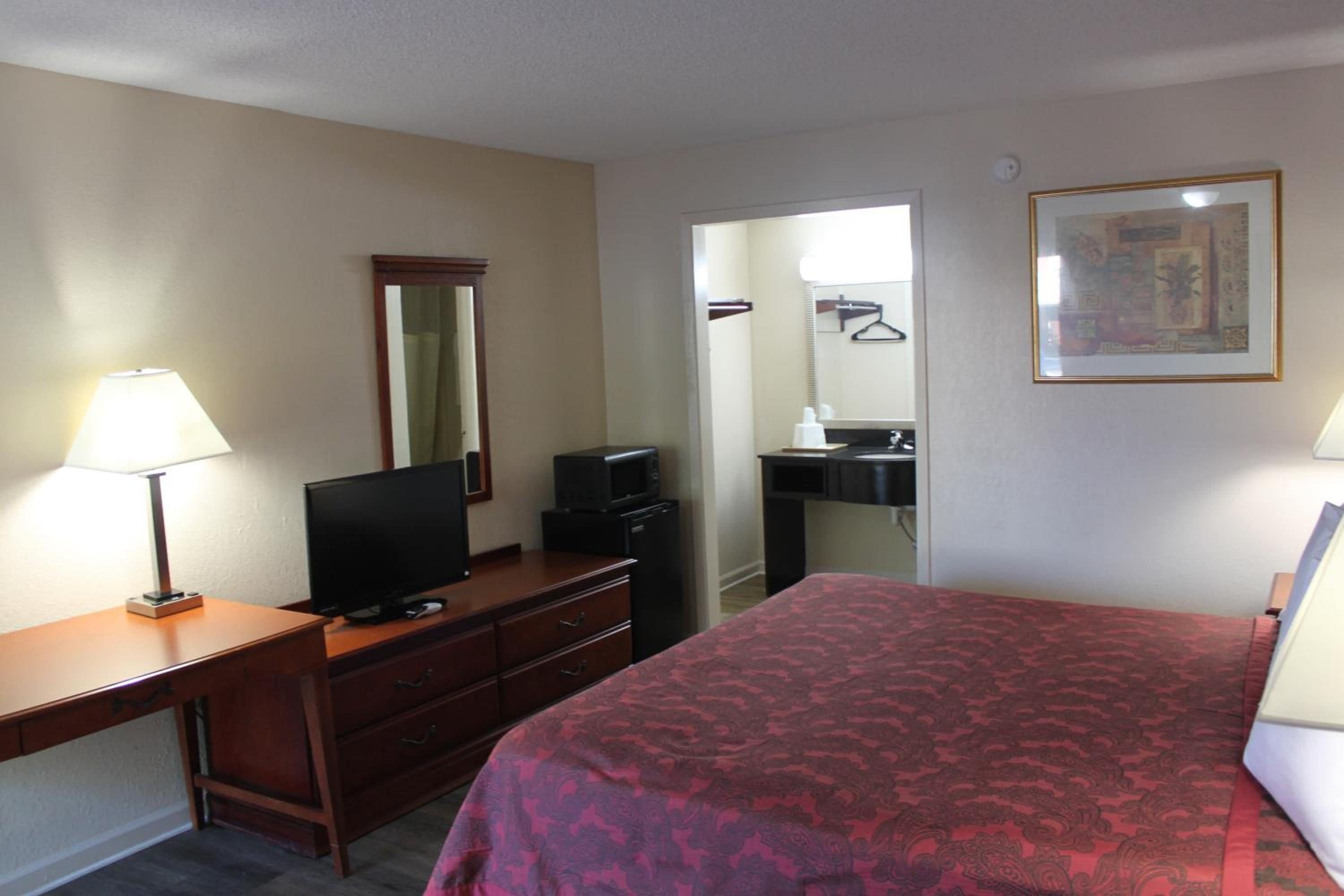 A hotel room with a bed, a desk with a television, a dresser, a mirror, a bathroom with a sink, and a framed picture on the wall.