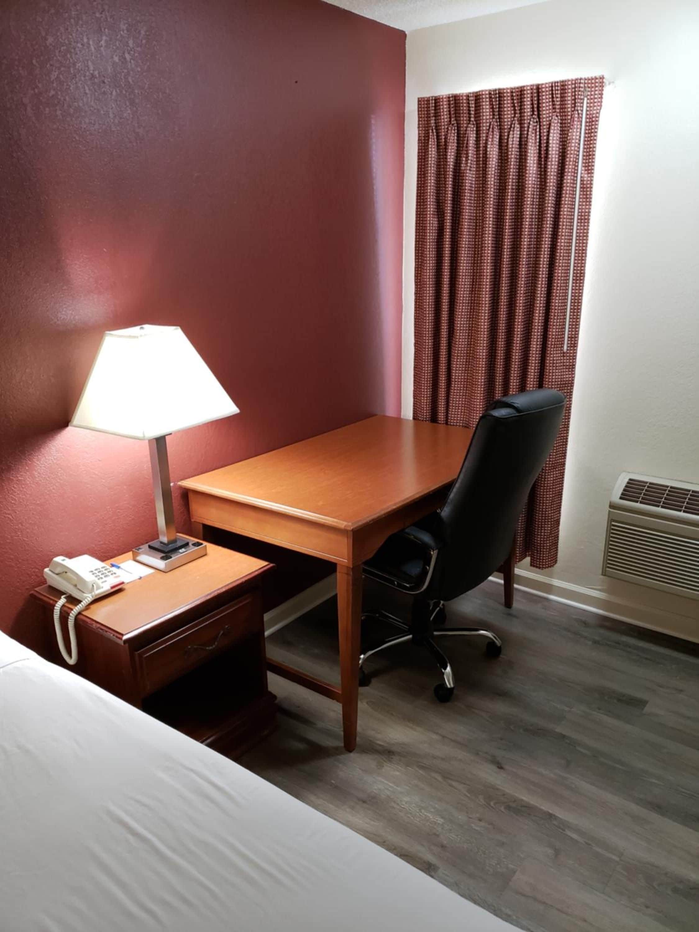 A hotel room with a wooden desk, a black office chair, a bedside table with a lamp, and a telephone.
