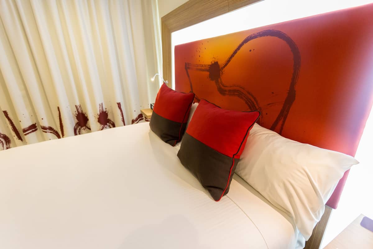 Una habitación de hotel con una cama grande, dos almohadas rojas y una blanca, un cabecero de madera y una ventana con persianas.