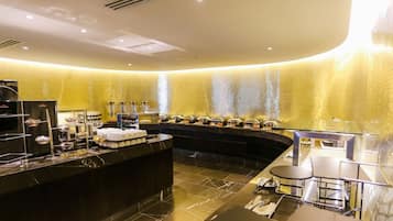 Een moderne hotellobby met een gebogen receptiebalie, een buffetgedeelte met food stations, en een zithoek met tafels en stoelen.