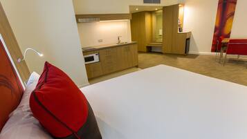 A hotel room with a bed, a red pillow, a small kitchenette, and a red chair.