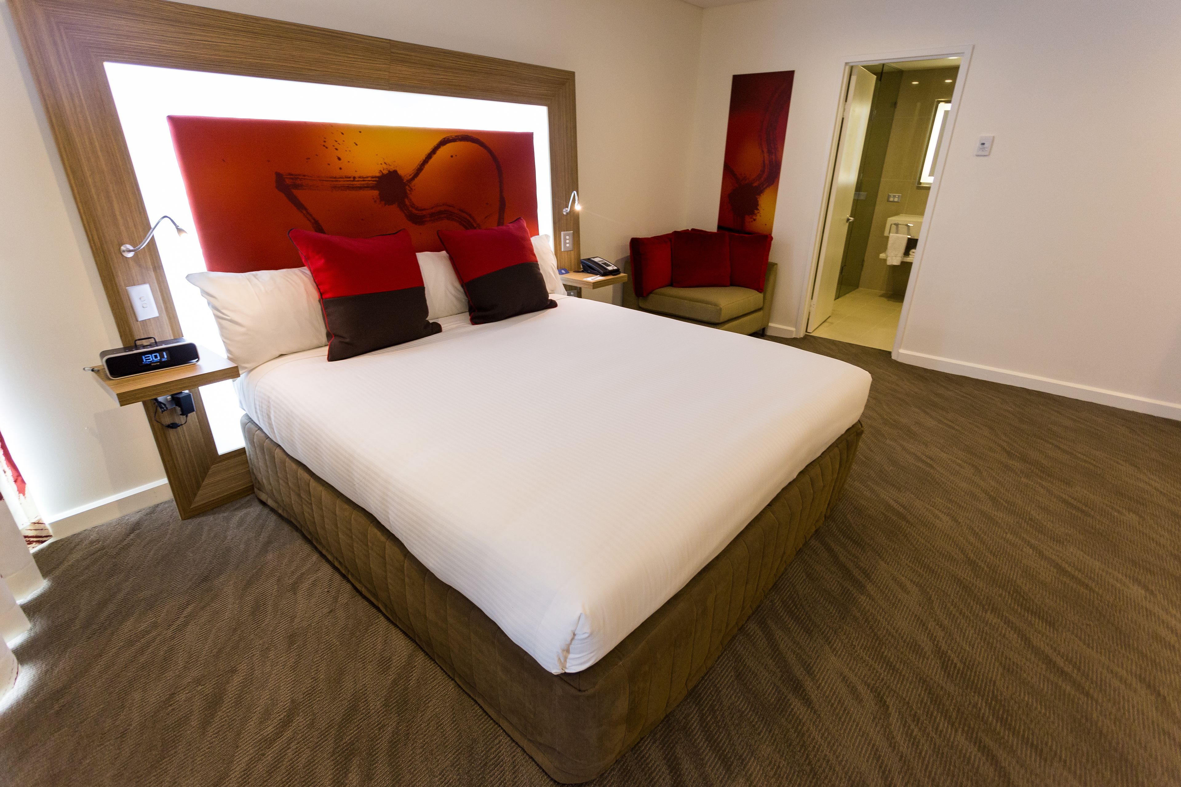 A hotel room with a large bed, a bedside table with a lamp, a chair, and a bathroom visible through an open door.