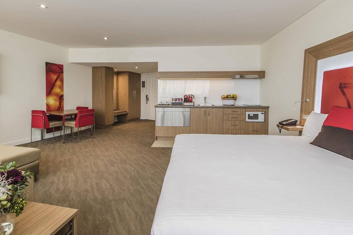 Habitación de hotel con una cama grande, un escritorio, una silla, una kitchenette pequeña y una zona de comedor con mesa y sillas.