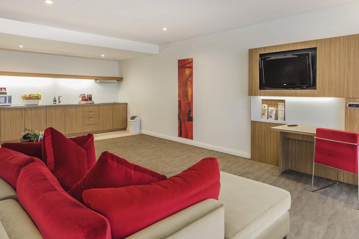 Una sala de estar moderna con un sofá beige, cojines rojos, un escritorio de madera, un televisor y una zona de cocina con armarios y un microondas.
