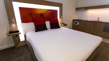 A hotel room with a large bed, a bedside table with a clock, a nightstand, a wooden headboard, a small desk with a telephone, and a kitchenette area.