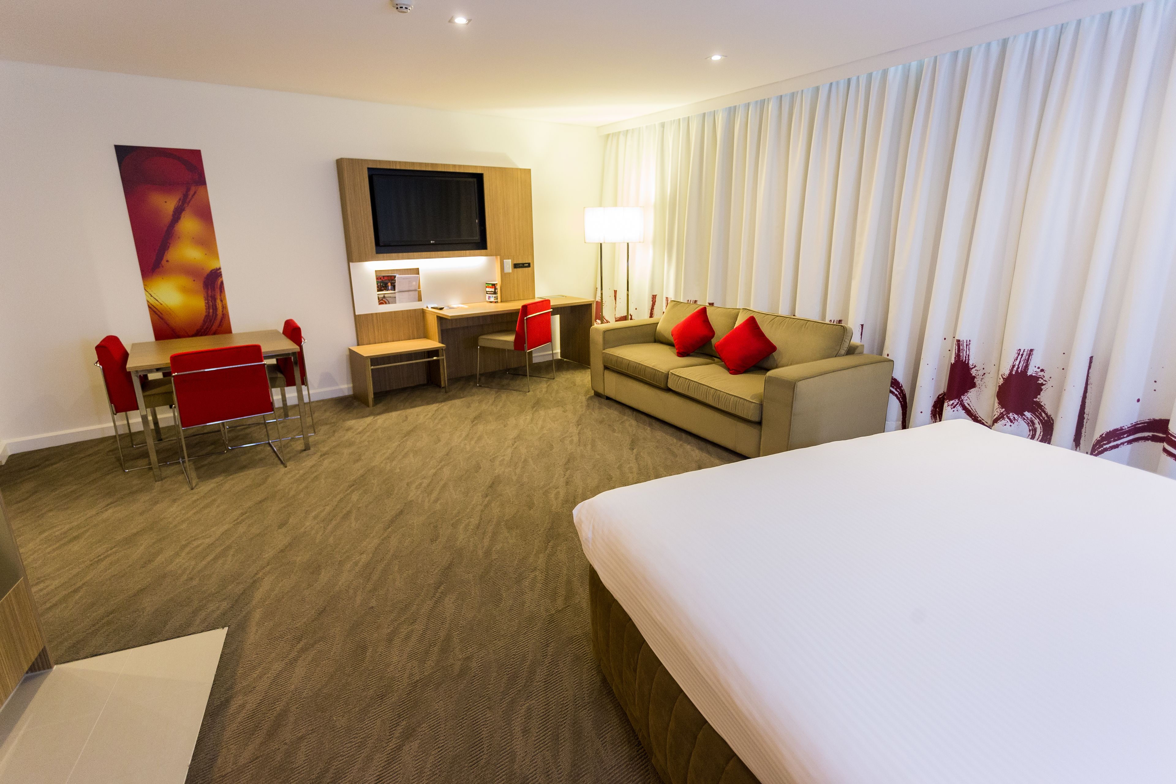 A hotel room with a bed, a desk with a chair, a sofa with red cushions, a TV, and a large window with curtains.
