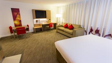 A hotel room with a bed, a desk with a chair, a sofa with red cushions, a TV, and a large window with curtains.