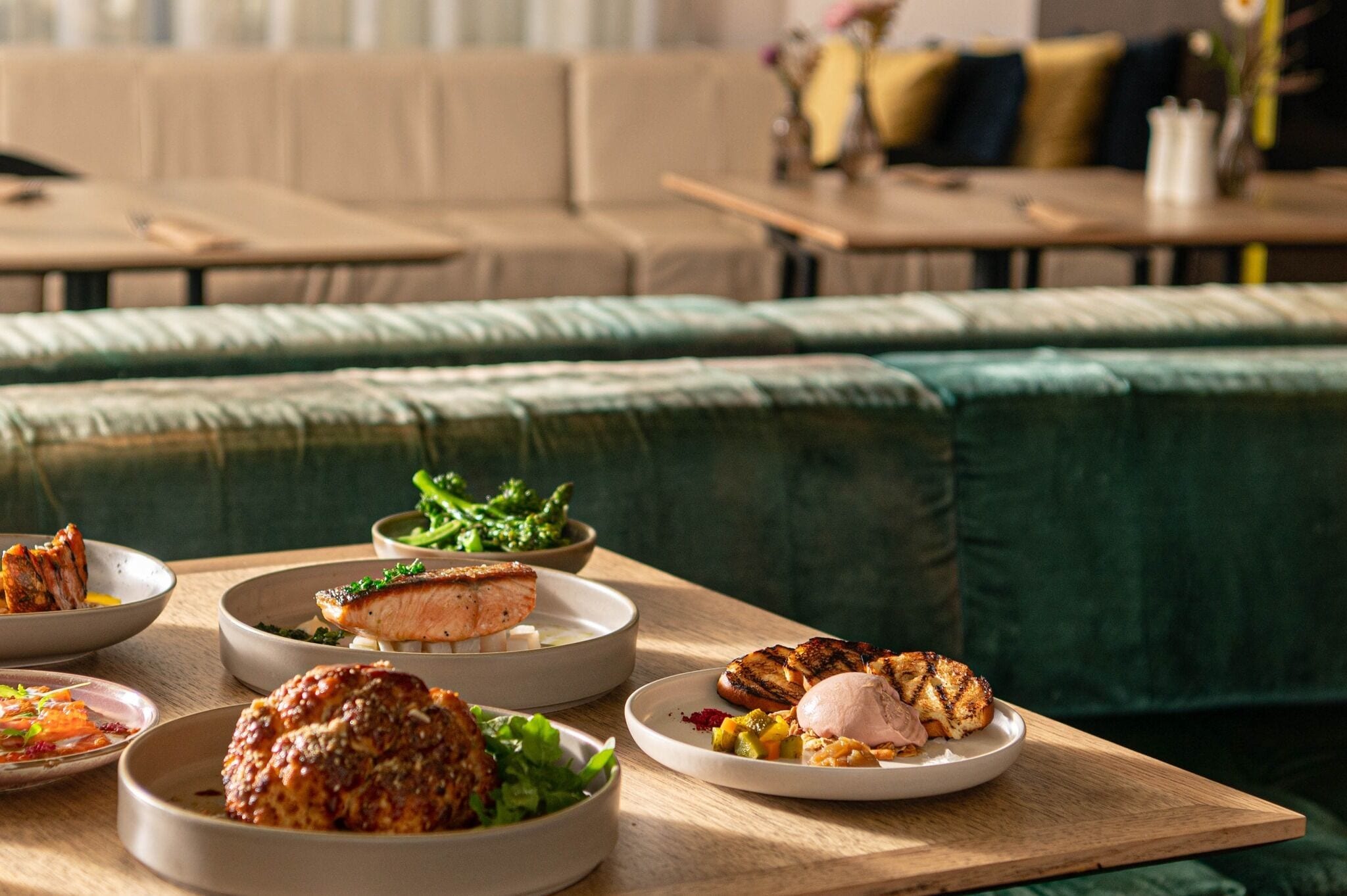 Zona pranzo con tavolo in legno apparecchiato con diversi piatti, tra cui carne alla griglia e verdure, in un ambiente moderno con un divano imbottito verde sullo sfondo.