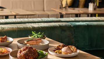 Zona pranzo con tavolo in legno apparecchiato con diversi piatti, tra cui carne alla griglia e verdure, in un ambiente moderno con un divano imbottito verde sullo sfondo.