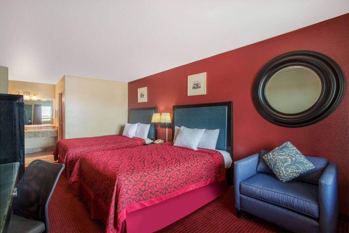 A hotel room with two beds, a chair, a mirror, and a desk with a lamp.