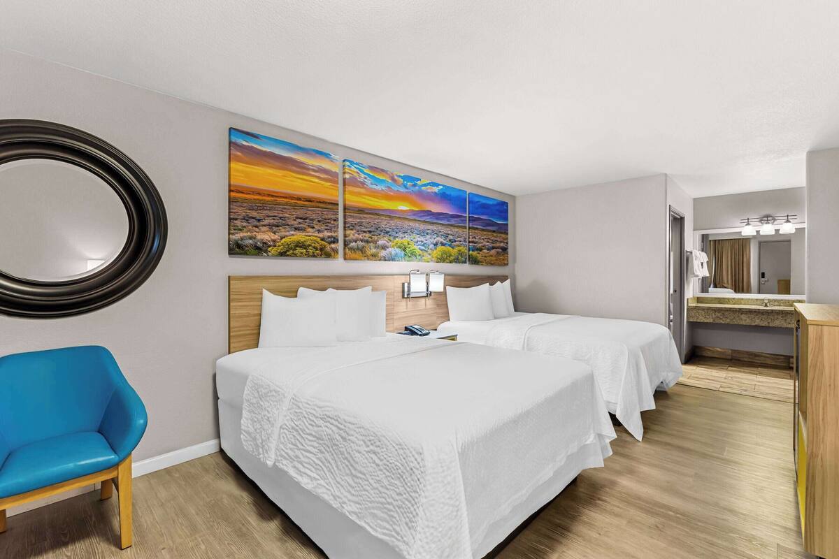 A hotel room with two beds, a blue chair, a large mirror, and a painting of a sunset.