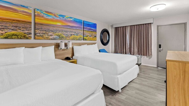 A hotel room with two beds, a wooden headboard, a desk, a chair, a window with curtains, and a wall with three paintings. - Days Inn by Wyndham Buckeye (Buckeye)
