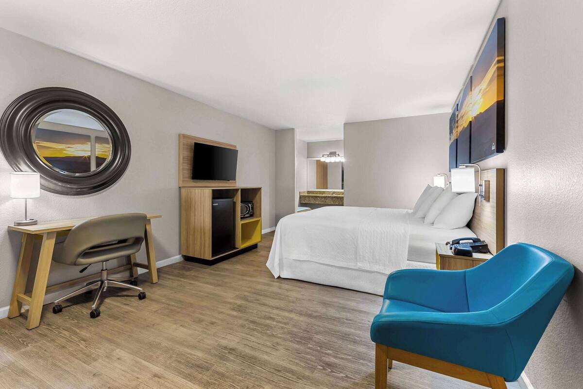 A hotel room with a bed, a desk with a chair, a TV, a mirror, and artwork on the walls.