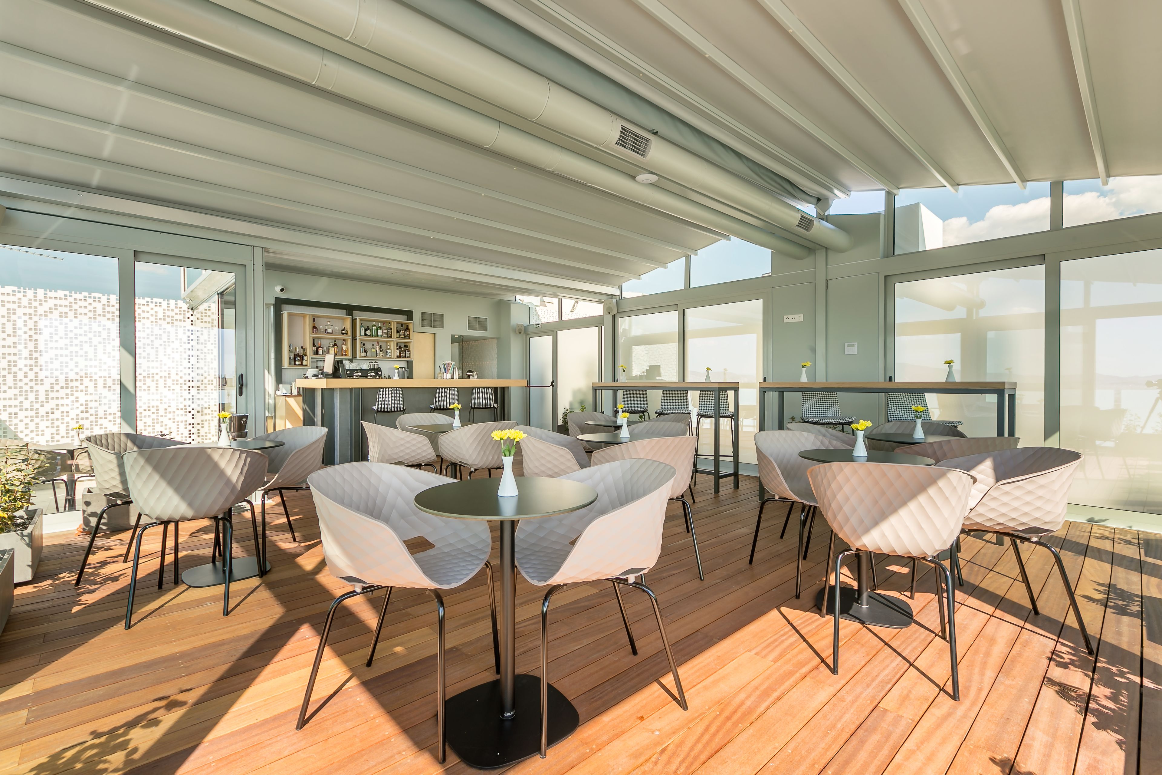 A spacious outdoor seating area with round tables and cushioned chairs, large windows offering views of the city, and a modern architectural design.