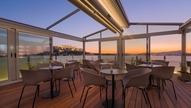 A rooftop terrace with tables and chairs, large windows, and a view of a cityscape at sunset.