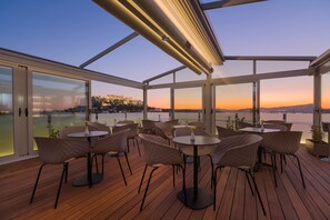 Terraço no telhado com mesas e cadeiras, janelas amplas e vista para o horizonte urbano ao pôr do sol.