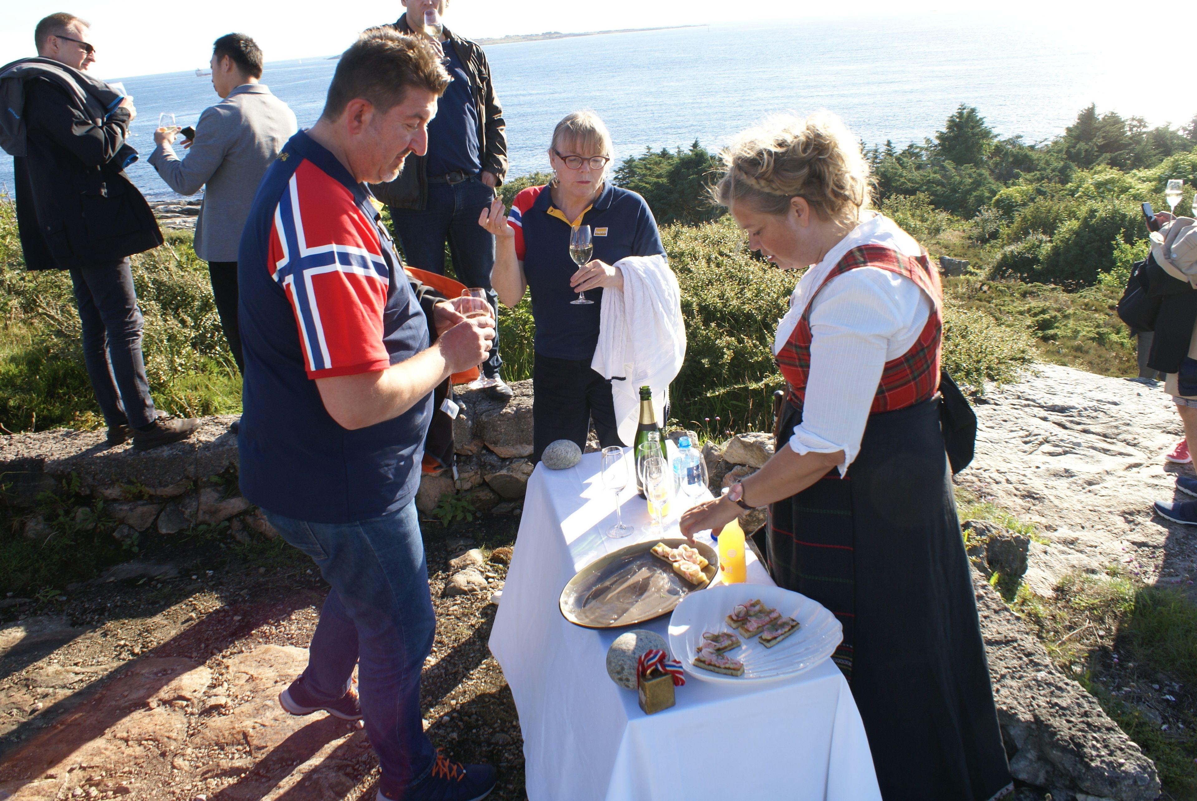 En gruppe mennesker ved et udendørs arrangement med et bord dækket til et måltid, med udsigt over et naturskønt kystlandskab.