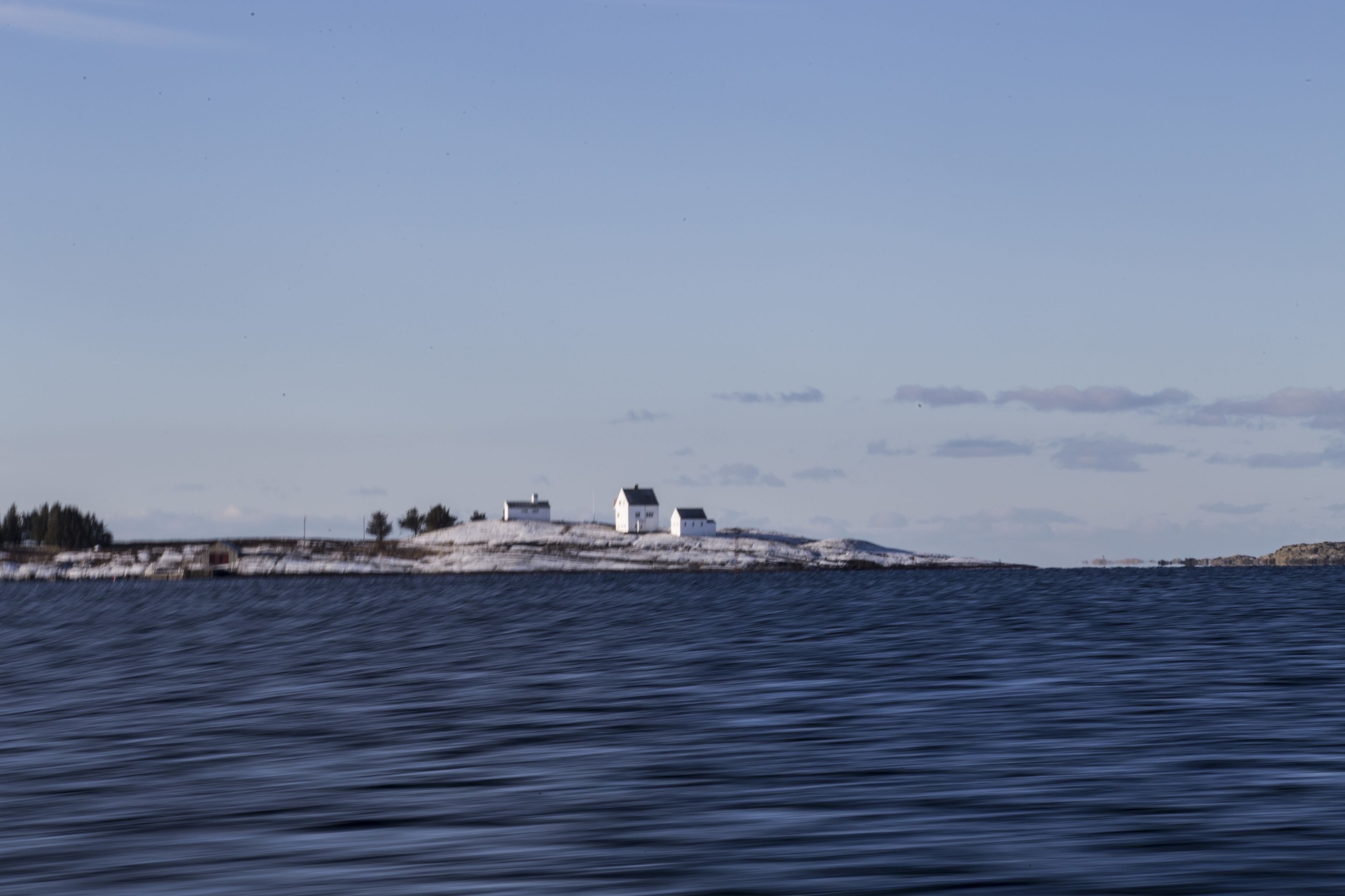 Escena costera con una pequeña isla que presenta algunos edificios, probablemente un faro u otra construcción similar, rodeada de aguas tranquilas.