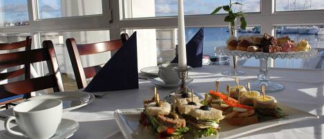 A table with a variety of sandwiches and a candle, set against a backdrop of a window with a view of boats.