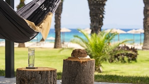 A hammock, a glass with a drink, and a book on a tree stump with a view of the ocean and beach umbrellas in the background. - Kassandra Palace Seaside Resort (Kassandra)