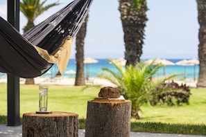 Un hamac, un verre avec une boisson, et un livre posé sur une souche d’arbre, avec en toile de fond l’océan et des parasols sur la plage.