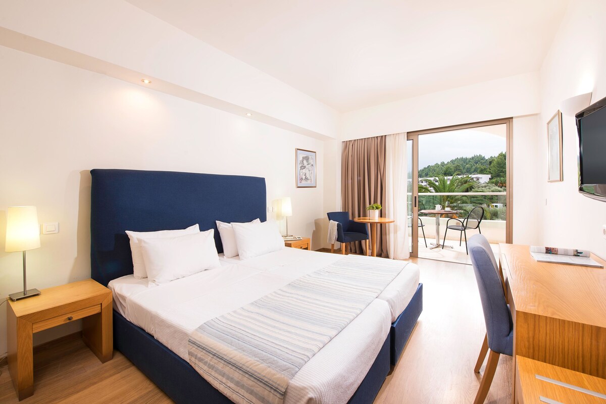 Ein Hotelzimmer mit einem großen Bett, einem Schreibtisch, einem Sessel, einem Fernseher und einem Balkon mit Blick.