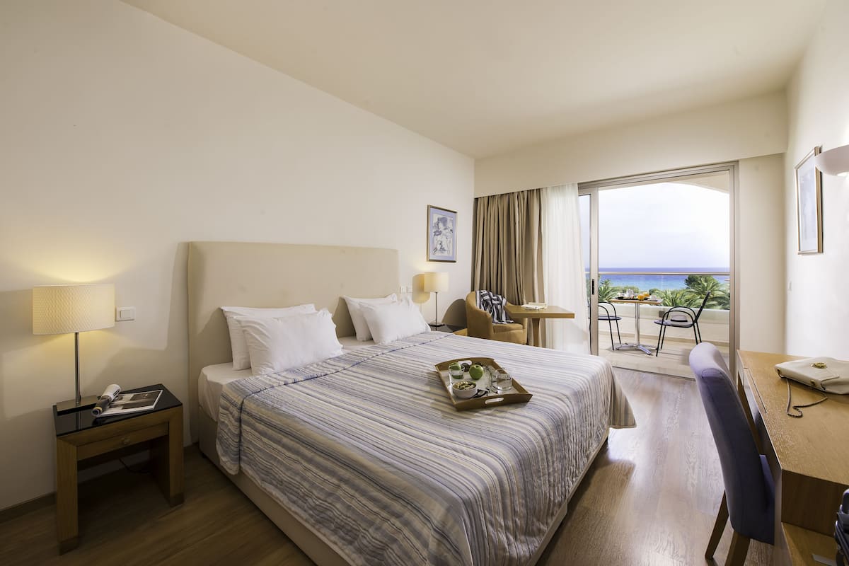 Une chambre d’hôtel avec un lit, un bureau, une chaise, une petite table, une lampe et une vue sur la mer.