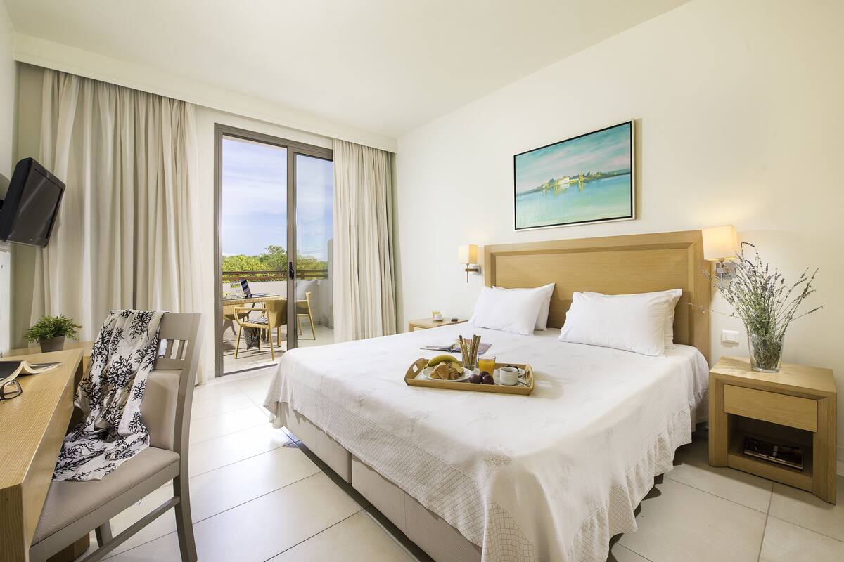 Une chambre d’hôtel comprenant un lit, un bureau, une chaise, une télévision, une fenêtre avec des rideaux, un tableau et un plateau avec des articles pour le petit-déjeuner.