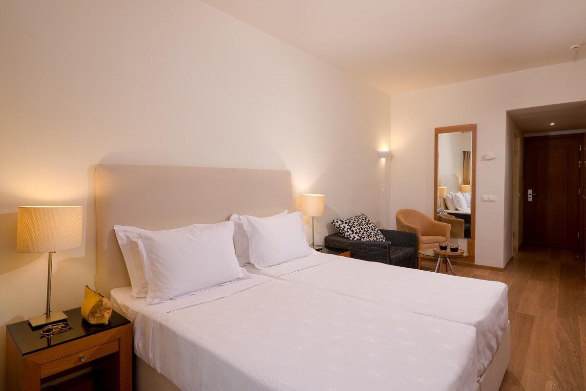 Ein Hotelzimmer mit einem großen Bett, einem Nachttisch mit Lampe, einem Sessel und einem kleinen Tisch mit Spiegel.