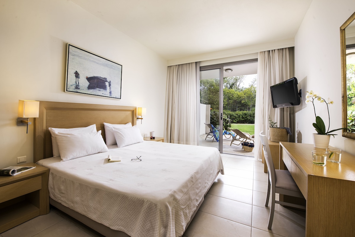 Ein Hotelzimmer mit einem großen Bett, einem Schreibtisch mit Stuhl, einem Fernseher und einem Balkon mit Blick auf Grünflächen.