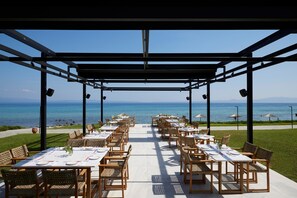 An outdoor dining area with tables and chairs arranged under a pergola, overlooking the sea. - Kassandra Palace Seaside Resort (Kassandra)