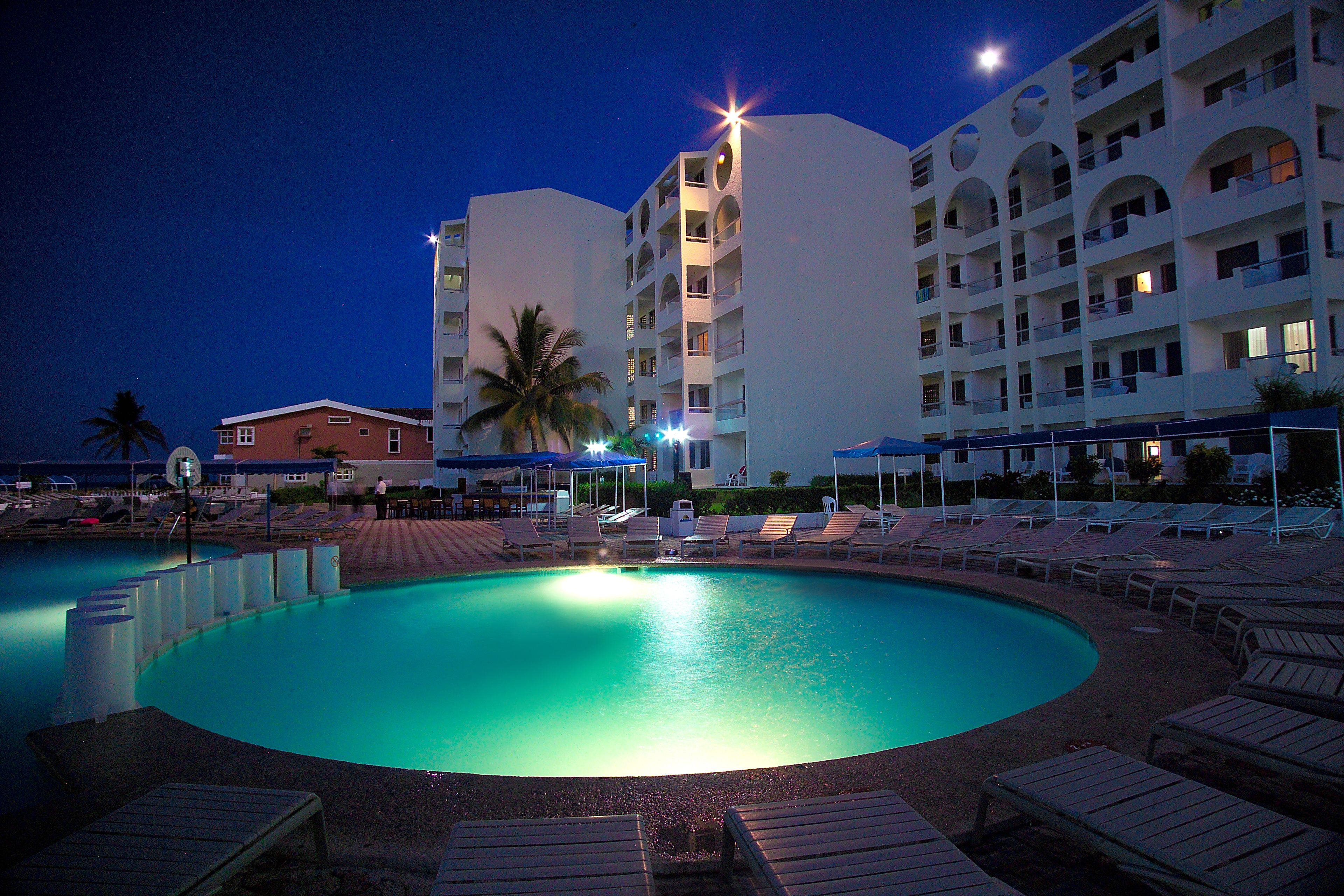 Zona de piscina con sillones reclinables y un edificio con varios balcones al fondo.