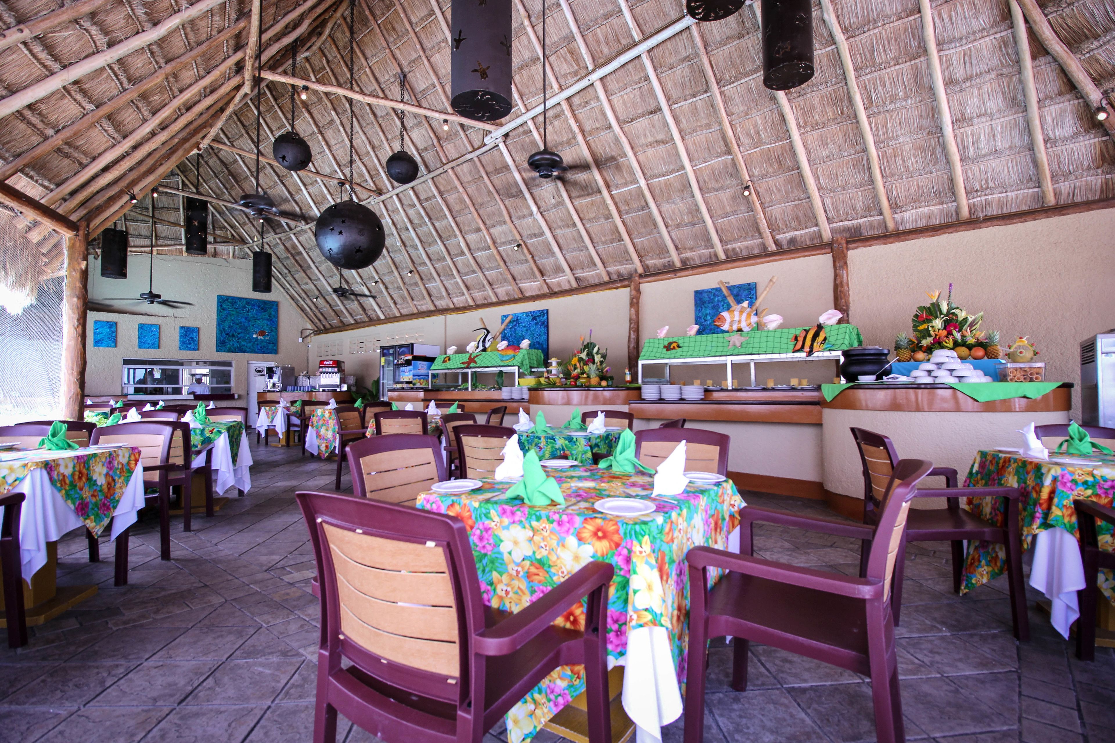 A restaurant with thatched roof, wooden tables, and chairs, featuring a kitchen in the background.